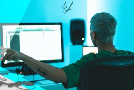 a man sits at a desk with a computer and microphone, engaged in an edm production course