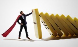 man in black suit with white cape stopping a stack of dominoes from falling