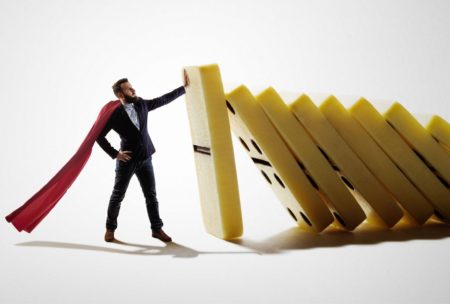 man in black suit with white cape stopping a stack of dominoes from falling