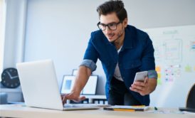 man typing on white laptop and holding silver mobile phone