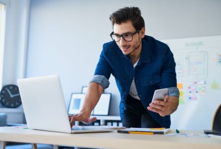 man typing on white laptop and holding silver mobile phone