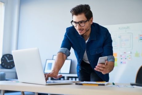 man typing on white laptop and holding silver mobile phone