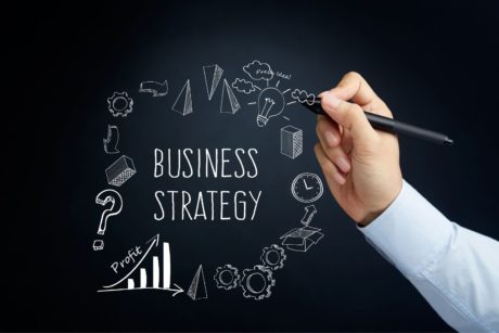 man writing the word business strategy and other buiness-related illustrations on a blackboard