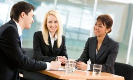 three people wearing suits sitting around a small table and talking to each other