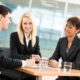 three people wearing suits sitting around a small table and talking to each other