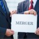 two men wearing suits holding white paper with the word merger