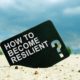 how to become resilient printed on black card lodged on white sand