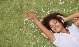 smiling woman in white top laying on grass listening to music on headphone