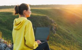 woman in yellow sweatshirt typing on laptop by the hills