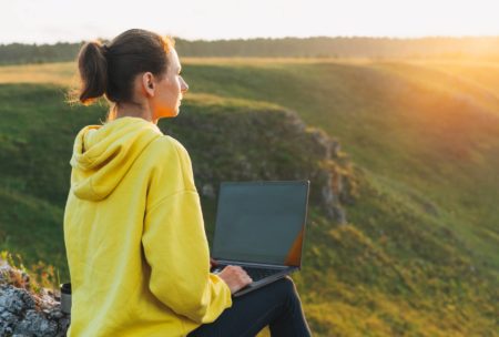 woman in yellow sweatshirt typing on laptop by the hills