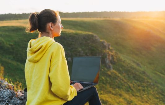 woman in yellow sweatshirt typing on laptop by the hills