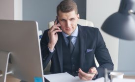 a man in a suit sits at his desk, engaged in a conversation on his cell phone, and email, focused on cold calling and cold emailing
