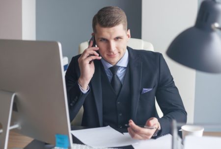 a man in a suit sits at his desk, engaged in a conversation on his cell phone, and email, focused on cold calling and cold emailing