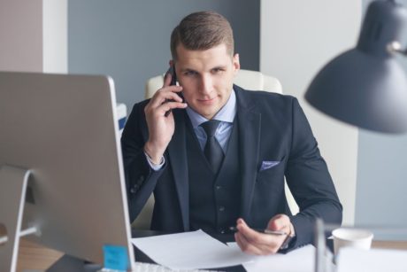a man in a suit sits at his desk, engaged in a conversation on his cell phone, and email, focused on cold calling and cold emailing