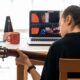 a woman plays an acoustic guitar while studying music theory guitar on her laptop