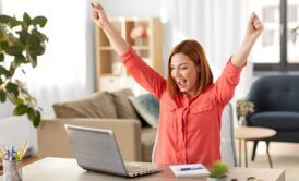 a woman at her desk, arms raised, engaged with her laptop, exemplifying effective time management strategies