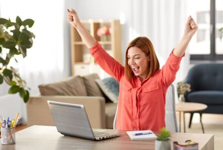 a woman at her desk, arms raised, engaged with her laptop, exemplifying effective time management strategies