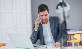 A man in a suit sits at a desk, using a laptop and talking on the phone. He is engaged in cold calling.