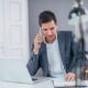 A man in a suit sits at a desk, using a laptop and talking on the phone. He is engaged in cold calling.