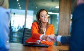 A woman in an orange suit shaking hands with another woman. Demonstrating professional etiquette during a job interview.