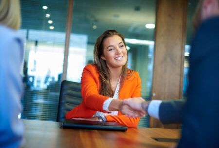 A woman in an orange suit shaking hands with another woman. Demonstrating professional etiquette during a job interview.