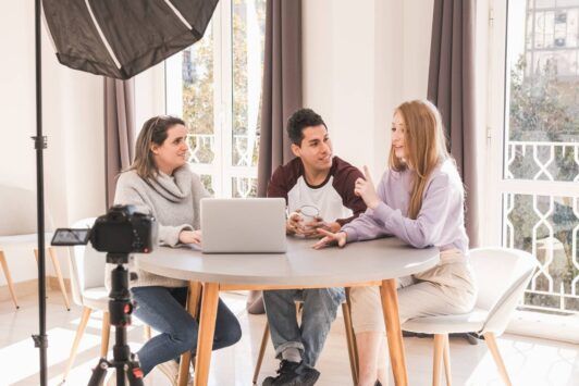 Three people at a table, using a laptop and camera for digital audience engagement.