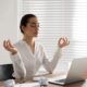 A woman sits at her desk, meditating with her laptop, embracing mindfulness in her work environment.