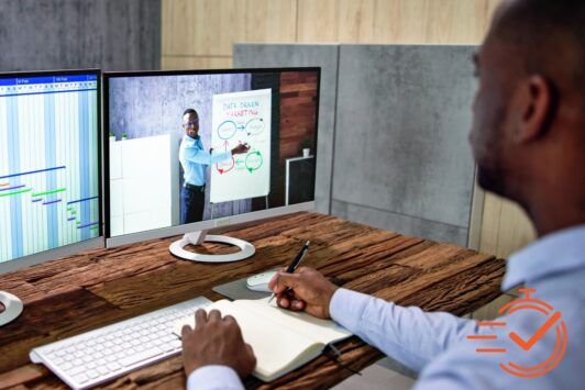 A man focused at his desk, using dual monitors to participate in remote training sessions via video.