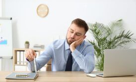 a man dressed in a shirt and tie works at a desk with a laptop, actively seeking strategies on how to overcome procrastination