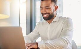 A smiling man sits at a table, working on his laptop, learning his microsoft skills in a productive atmosphere.