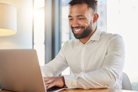 A smiling man sits at a table, working on his laptop, learning his microsoft skills in a productive atmosphere.