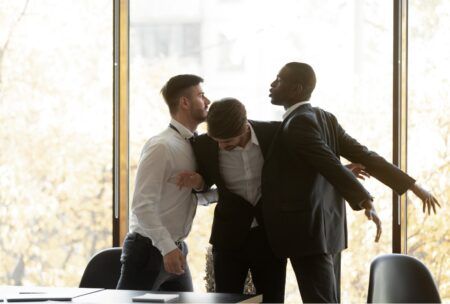 Three men in suits are engaged in a physical altercation inside an office setting, highlighting the need to prevent workplace violence.