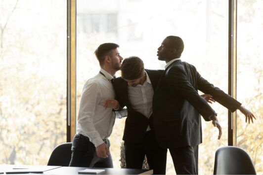 Three men in suits are engaged in a physical altercation inside an office setting, highlighting the need to prevent workplace violence.