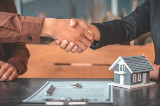 two people shaking hands over a house model, symbolizing a discussion on buying vs renting a home