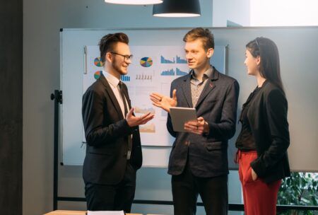 three business professionals collaborate around a table, reviewing a tablet related to project management courses
