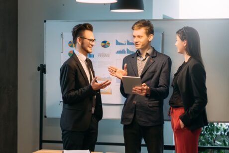 three business professionals collaborate around a table, reviewing a tablet related to project management courses