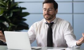 a man dressed in a shirt and tie sits at a desk, hands open, engaged with a laptop, reflecting the concept of peaceful chaos