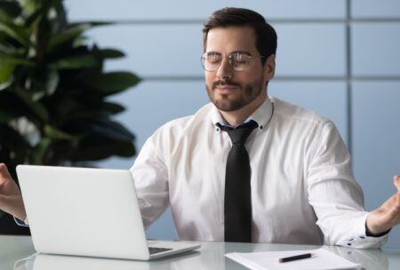 a man dressed in a shirt and tie sits at a desk, hands open, engaged with a laptop, reflecting the concept of peaceful chaos