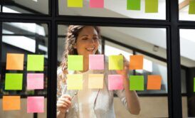 a woman observes a window filled with sticky notes, illustrating her involvement in a scrum master course