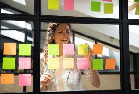 a woman observes a window filled with sticky notes, illustrating her involvement in a scrum master course