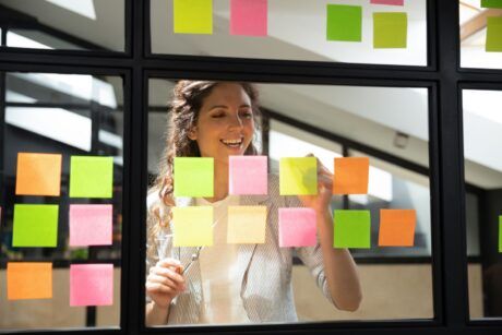 a woman observes a window filled with sticky notes, illustrating her involvement in a scrum master course